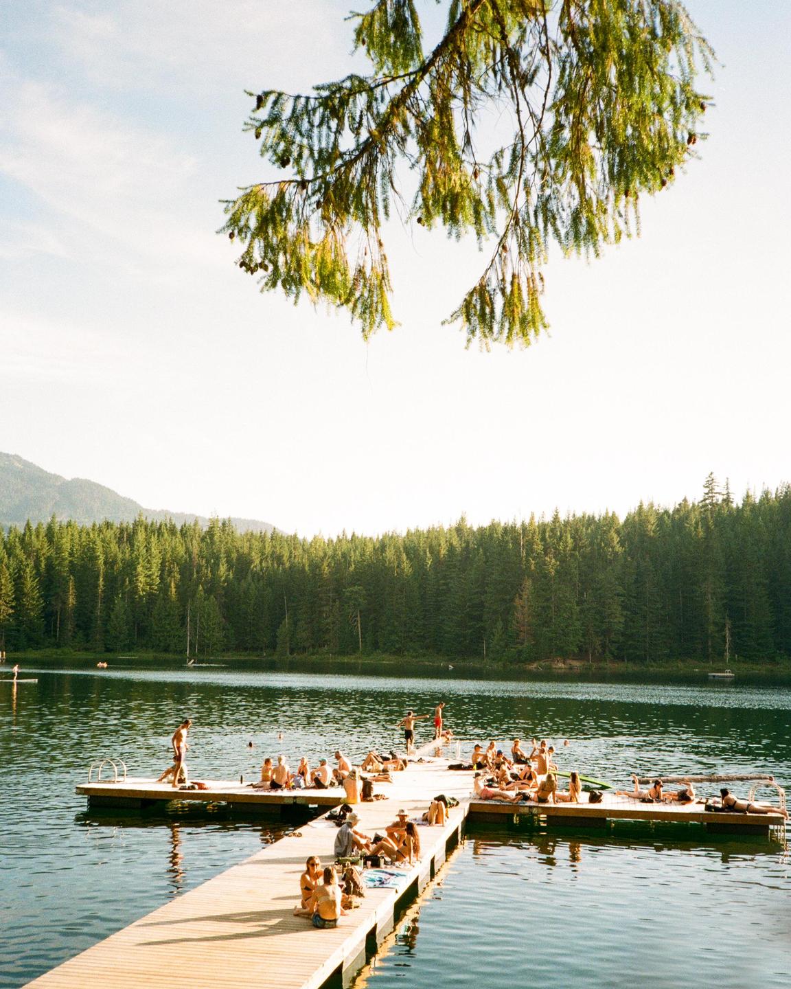 A group of people are relaxing on a wooden dock on a lake surrounded by a forest.