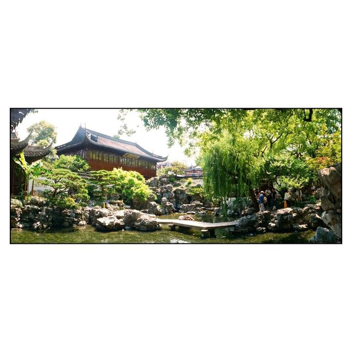 A traditional Chinese building with a dark roof stands behind a pond with rocks and lush green trees.