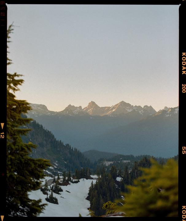 A mountain range with snow-covered slopes and evergreen trees is visible under a clear sky.