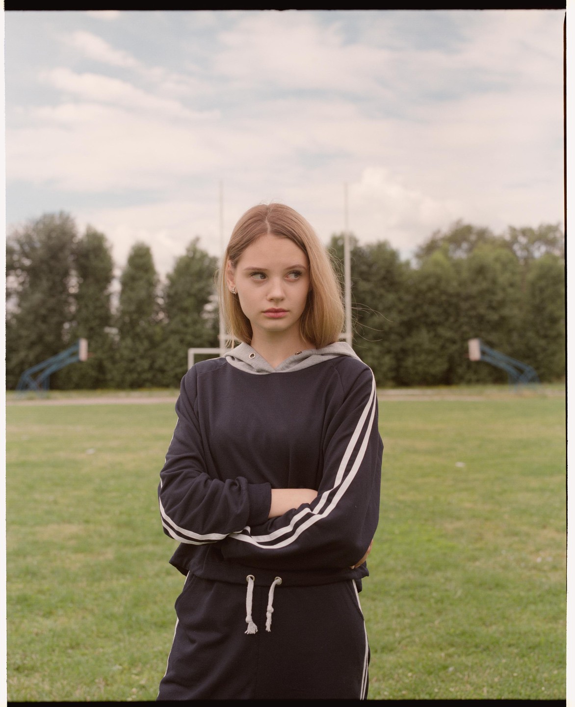 A young woman stands with her arms crossed on a grassy field with trees and sports equipment in the background.