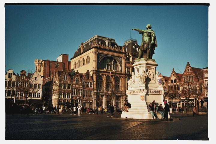 A statue of a man pointing stands in a city square surrounded by historic buildings and people.