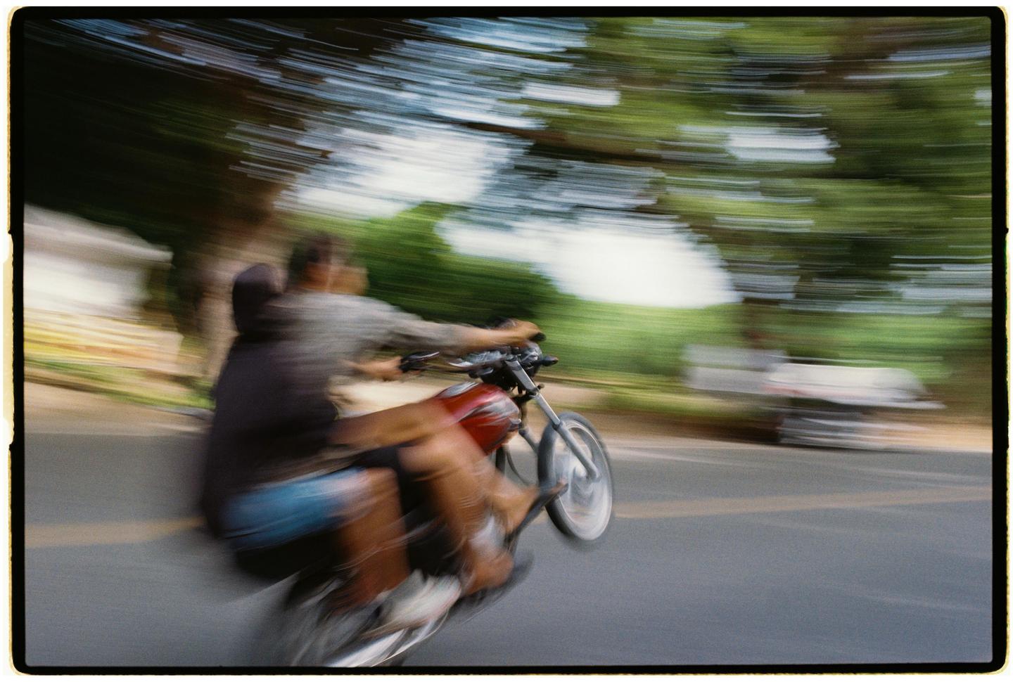 Two people ride a motorcycle on a road with blurred trees and a vehicle in the background.