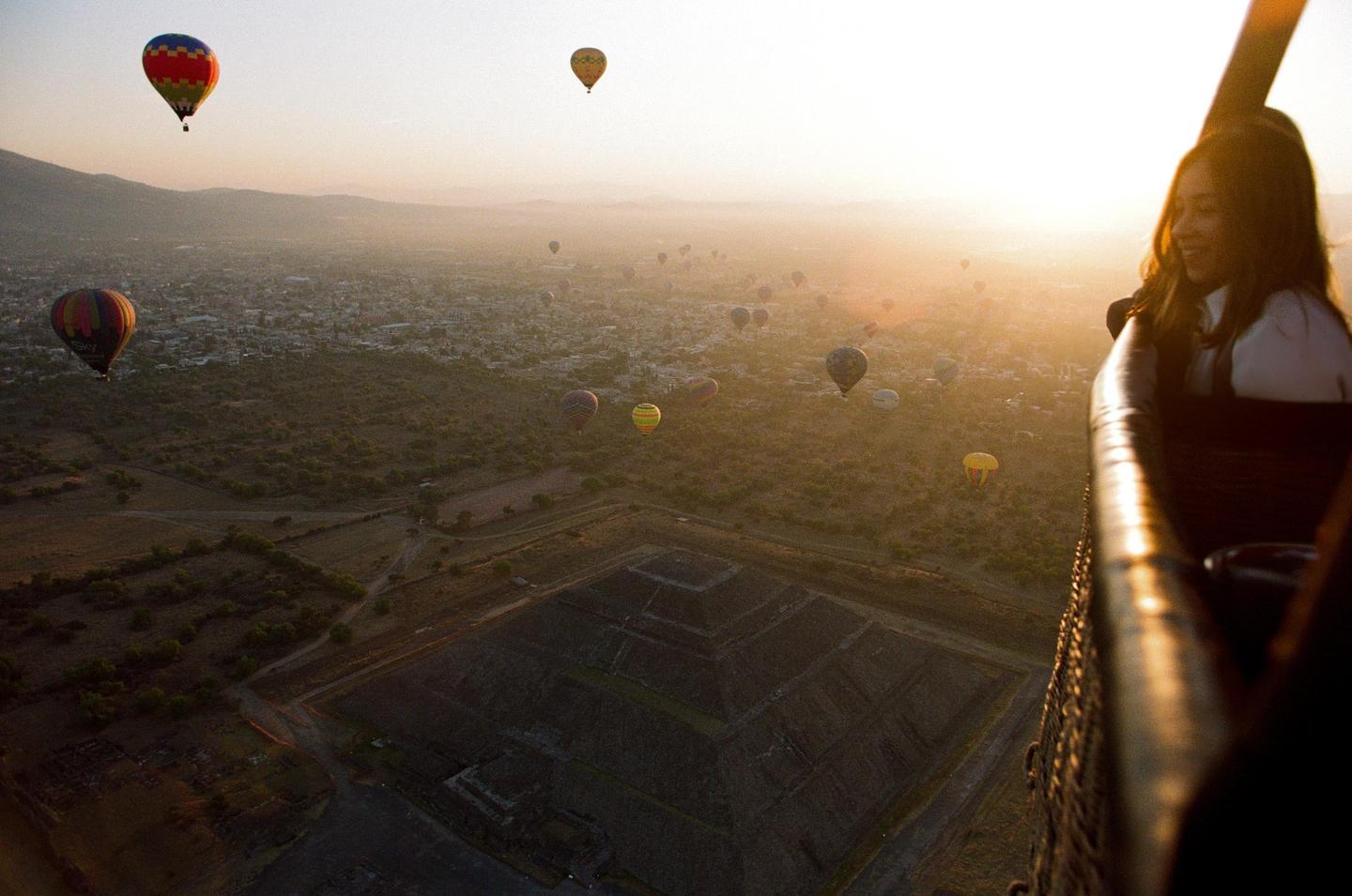 A woman looks out from a hot air balloon over a landscape with a pyramid and other hot air balloons at sunset.