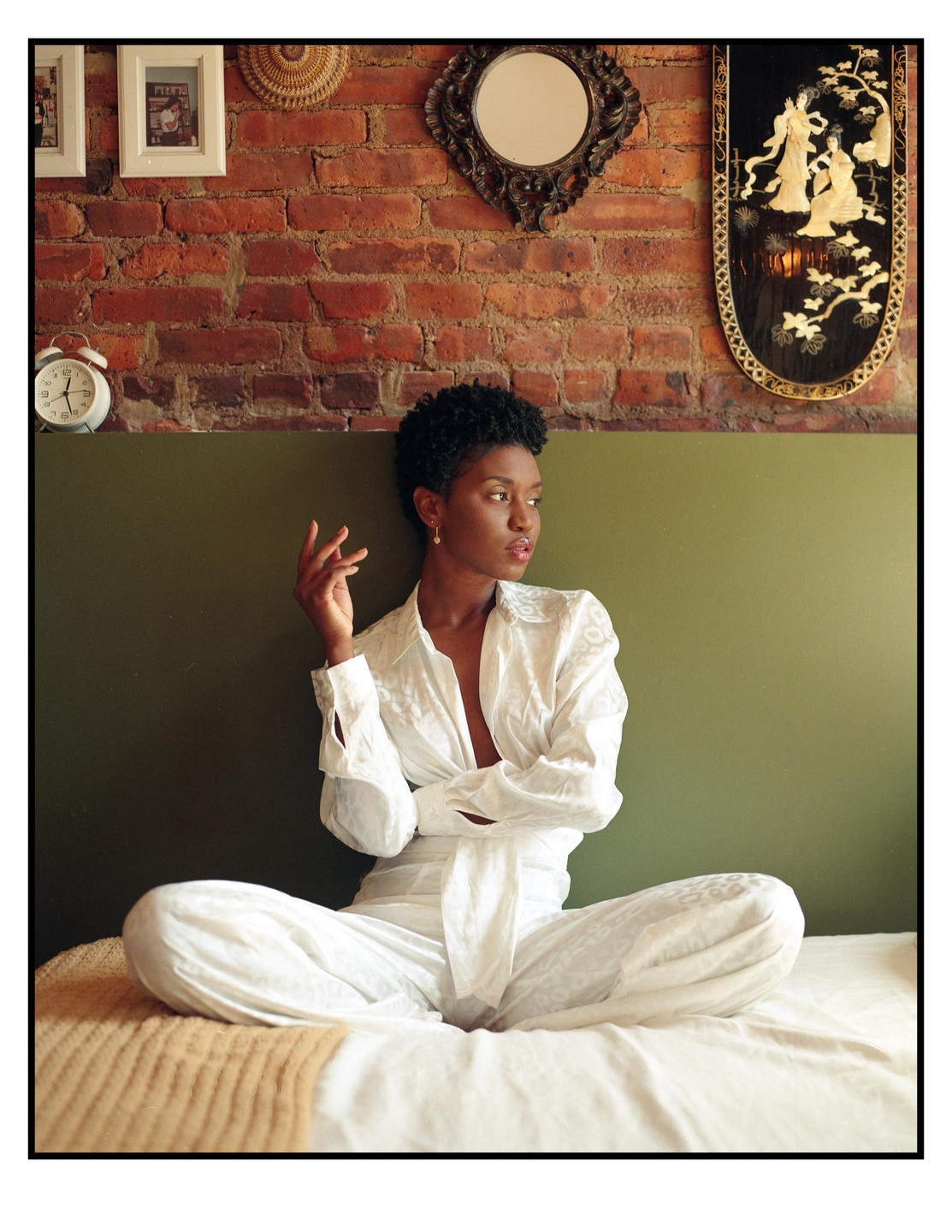 A woman in white pajamas sits cross-legged on a bed in a room with a brick wall and decorative items.