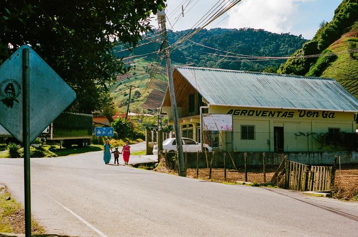 Two women and a child walk along a road past a building with a metal roof and a sign that reads "AGROVENTAS Don Ga" in a mountainous landscape.