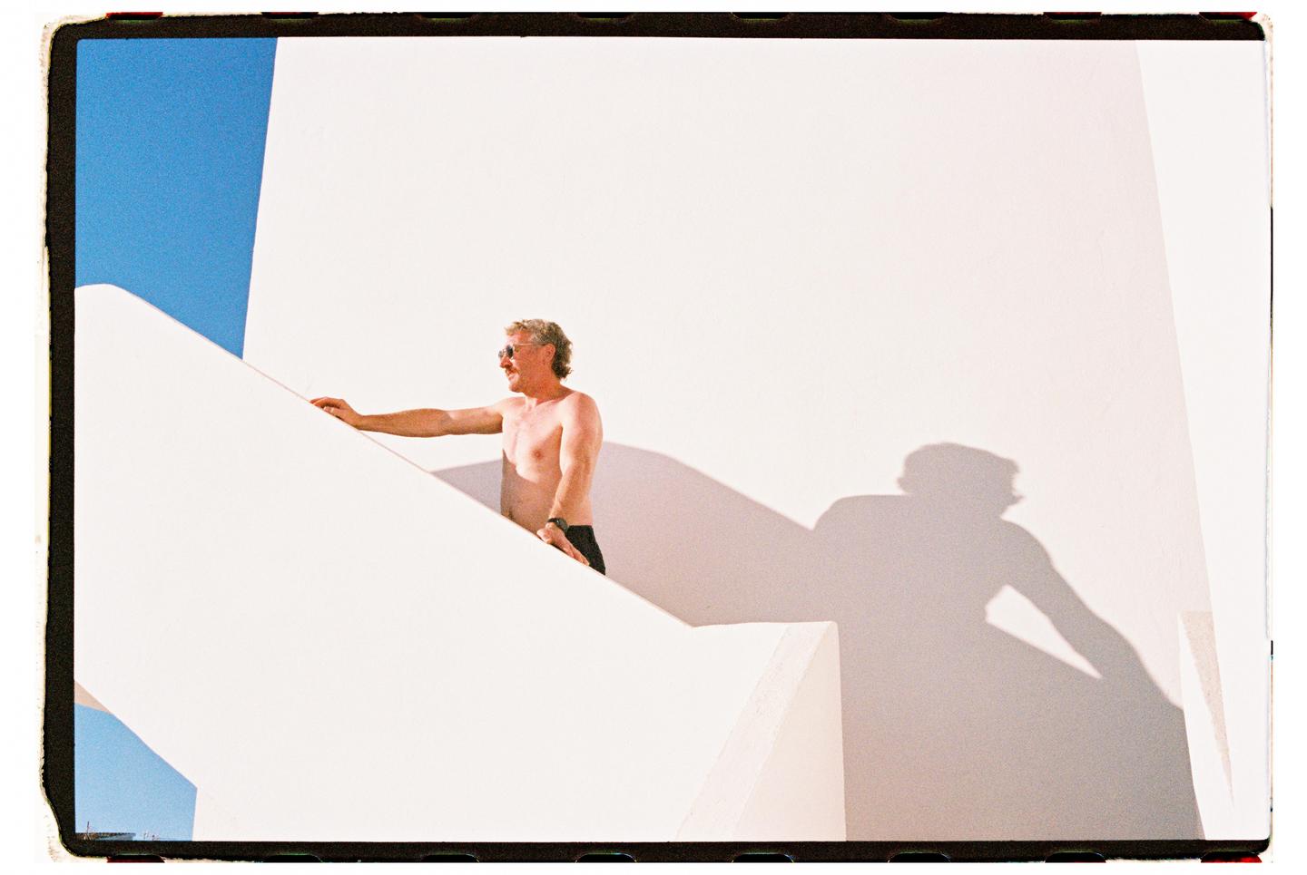 A man in sunglasses and swim trunks stands on a white architectural structure with a blue sky in the background.