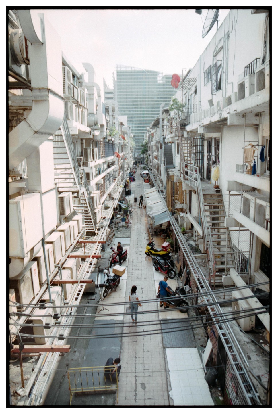 A narrow alleyway is lined with buildings and air conditioning units, with people and motorbikes on the ground.