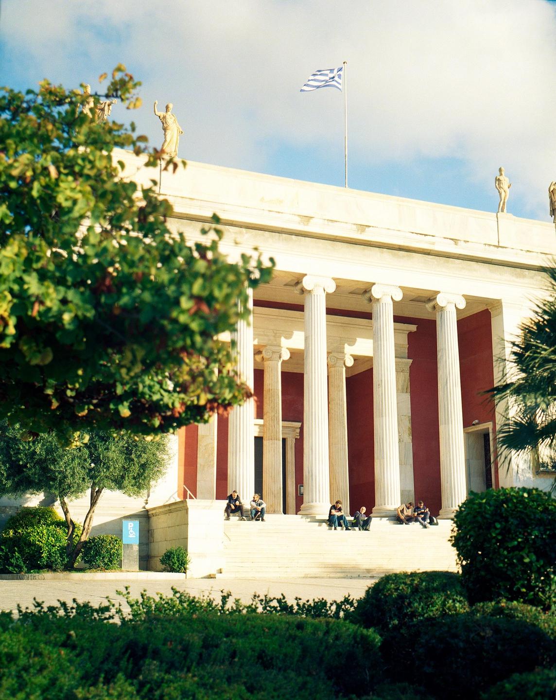 A group of people sit on the steps of a large building with columns and a Greek flag flying from the roof.