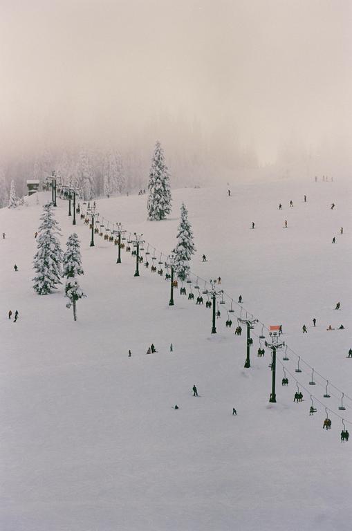 A ski slope with a chairlift and many skiers is covered in snow, with snow-covered trees and a hazy sky in the background.