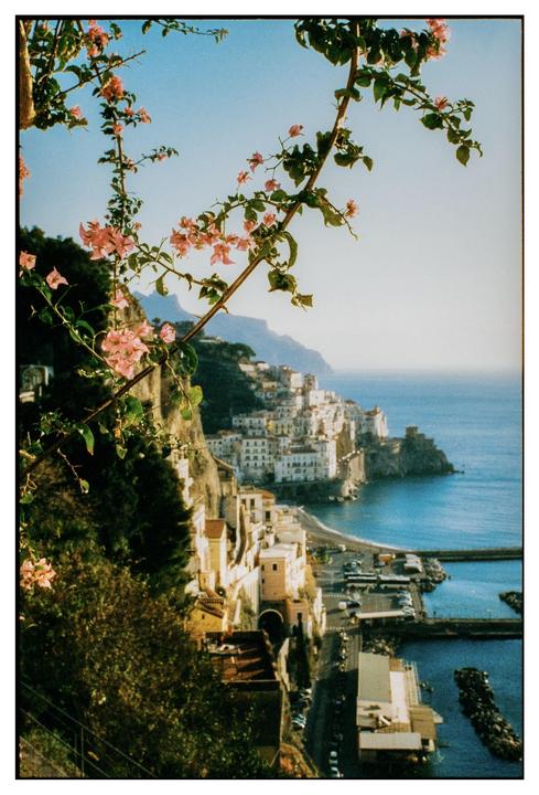 Pink bougainvillea flowers frame a view of a coastal town built into a cliffside overlooking the sea.