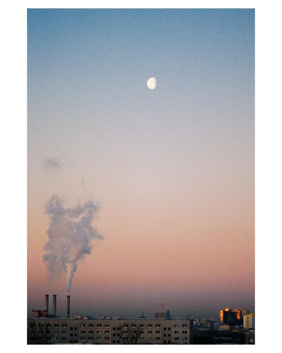 A full moon hangs in a gradient sky above a city skyline with smoke rising from industrial chimneys.