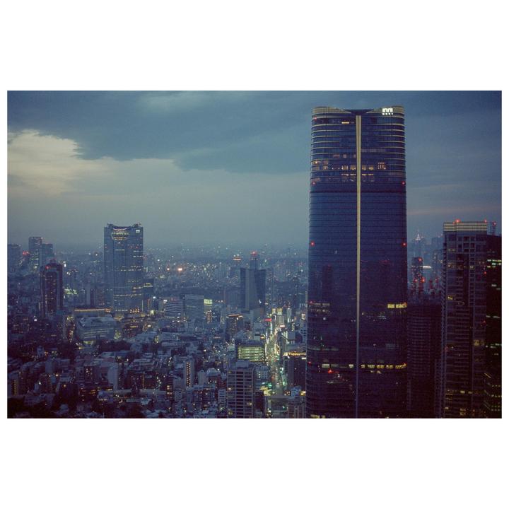 A tall, modern skyscraper with illuminated windows stands prominently against a city skyline at dusk.