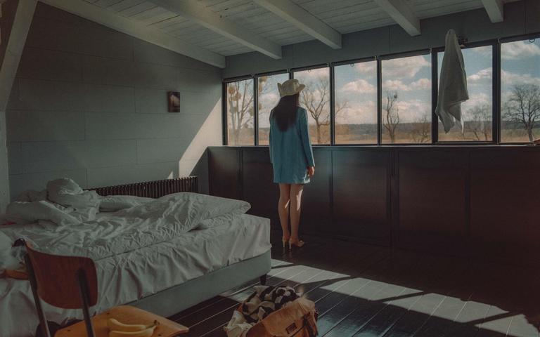 A woman in a hat and blue dress stands looking out a window in a bedroom with an unmade bed and sunlight streaming through the window.