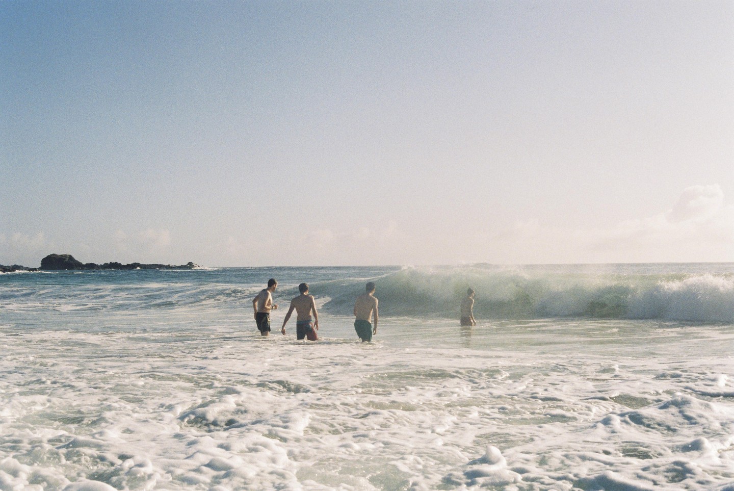 Four men stand in the ocean as a wave breaks behind them.
