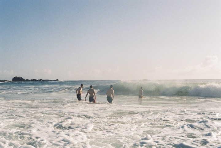 Four men stand in the ocean as a wave breaks behind them.