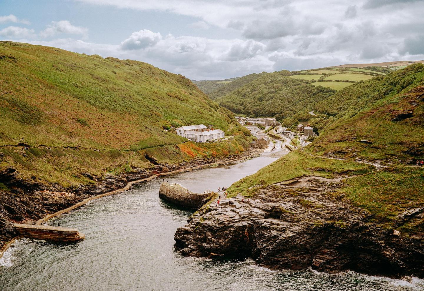 A small village with white buildings is nestled in a valley between green hills, with a river flowing through it and out to the sea.