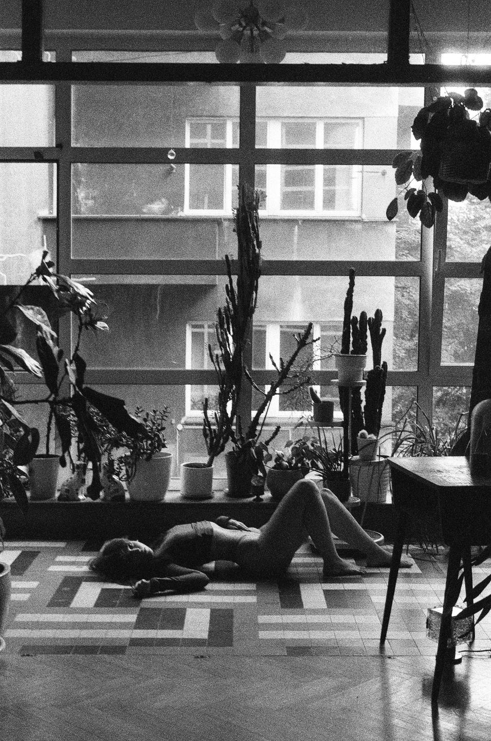 A woman lies on a patterned floor in front of a window with many potted plants and a view of a building.