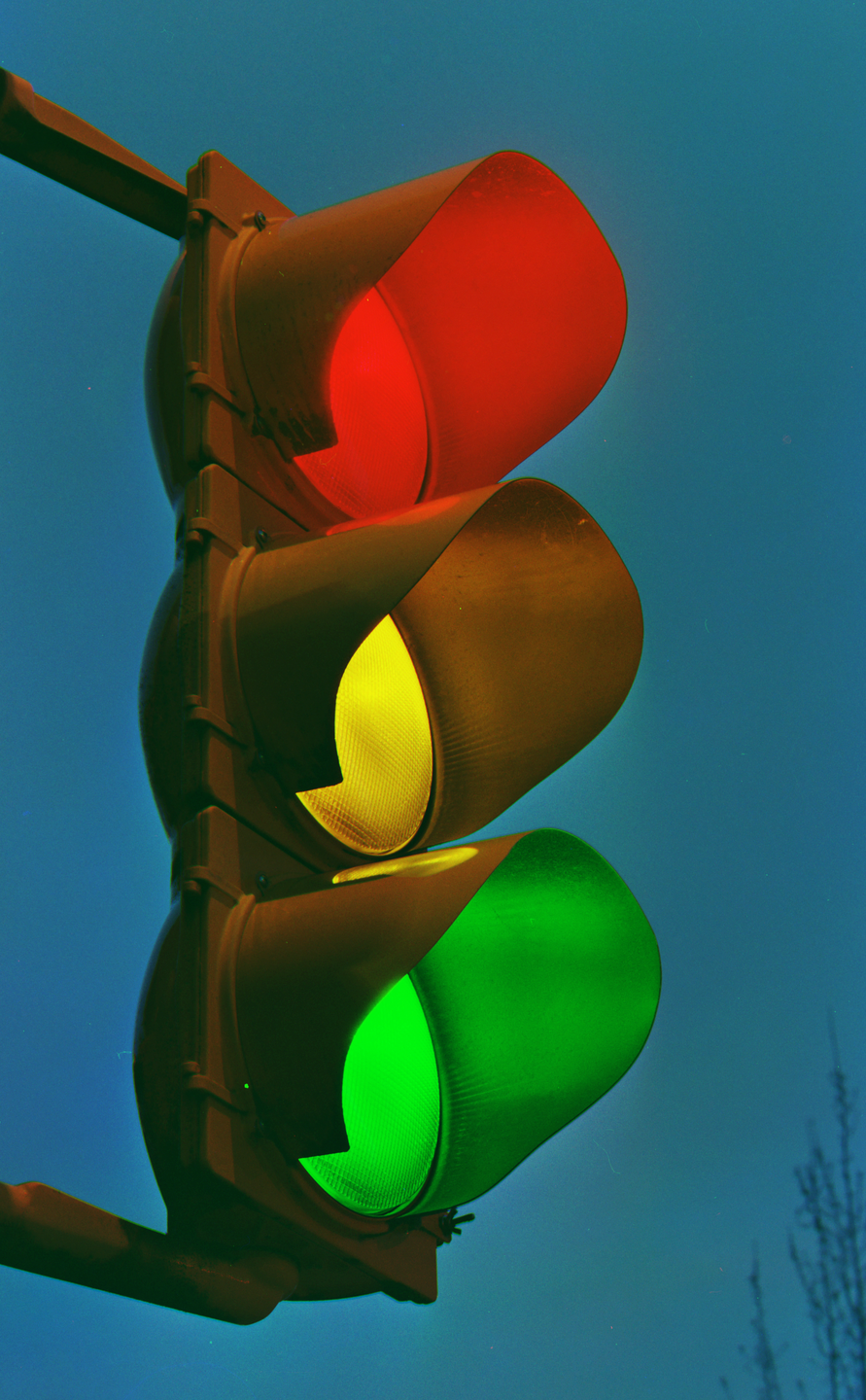 A traffic light with red, yellow, and green lights illuminated is viewed from below against a blue sky.