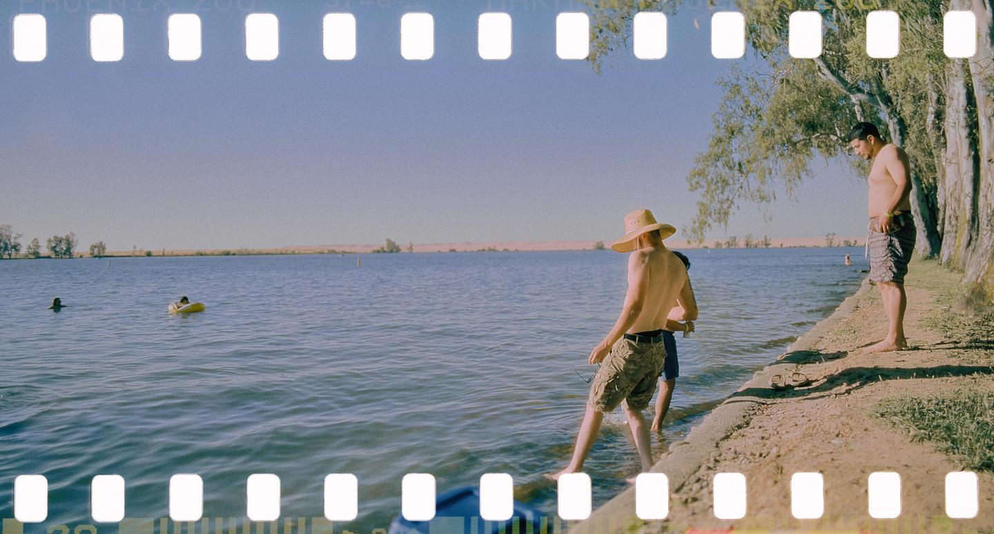 Three men are by a lake, with two in the water and one standing on the shore.