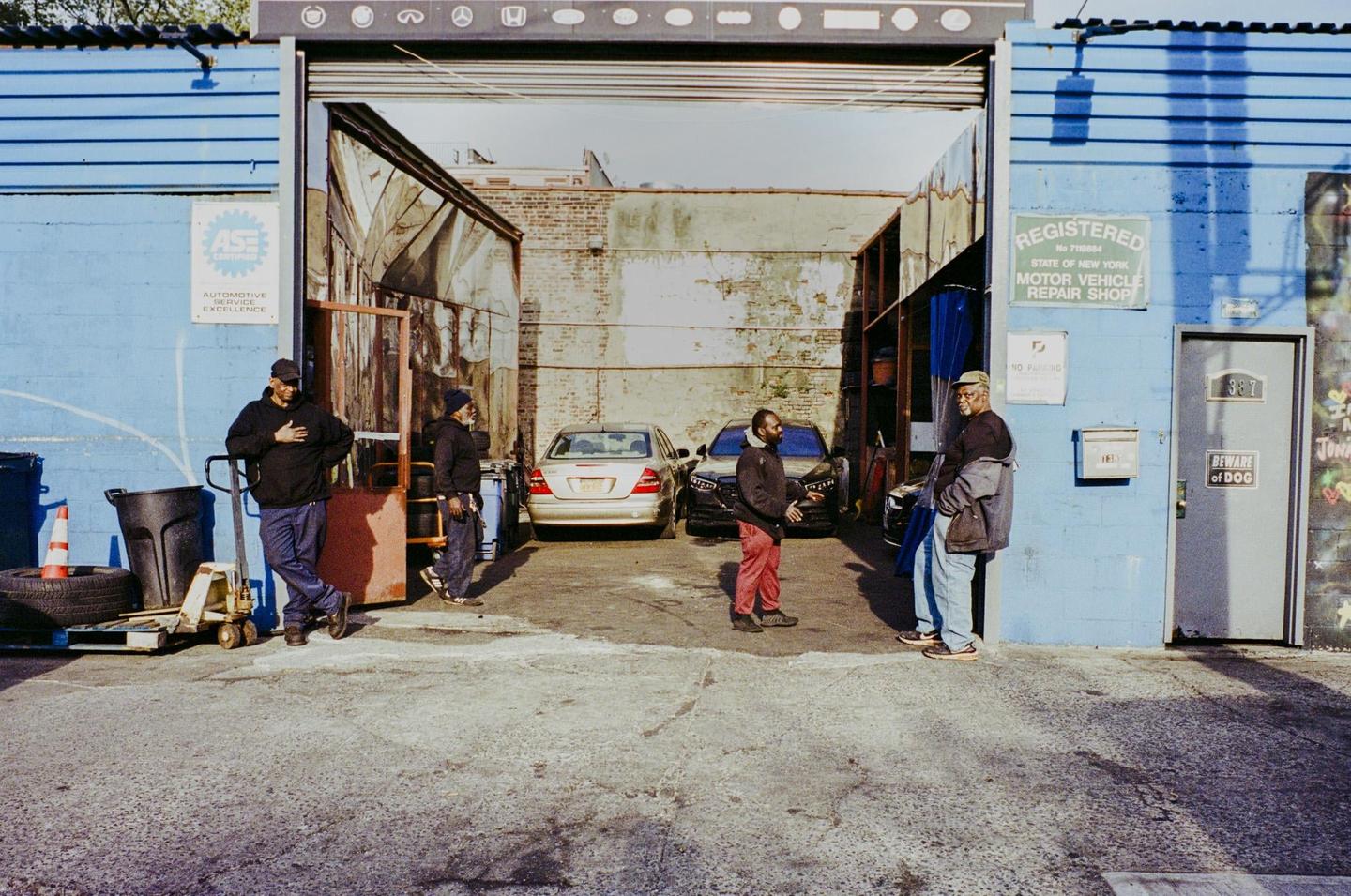 Four men are standing in and around an automotive repair shop with two cars parked inside.