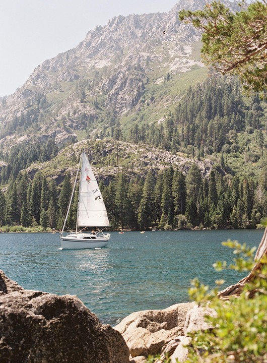 A sailboat with a white sail floats on a lake surrounded by mountains and pine trees.