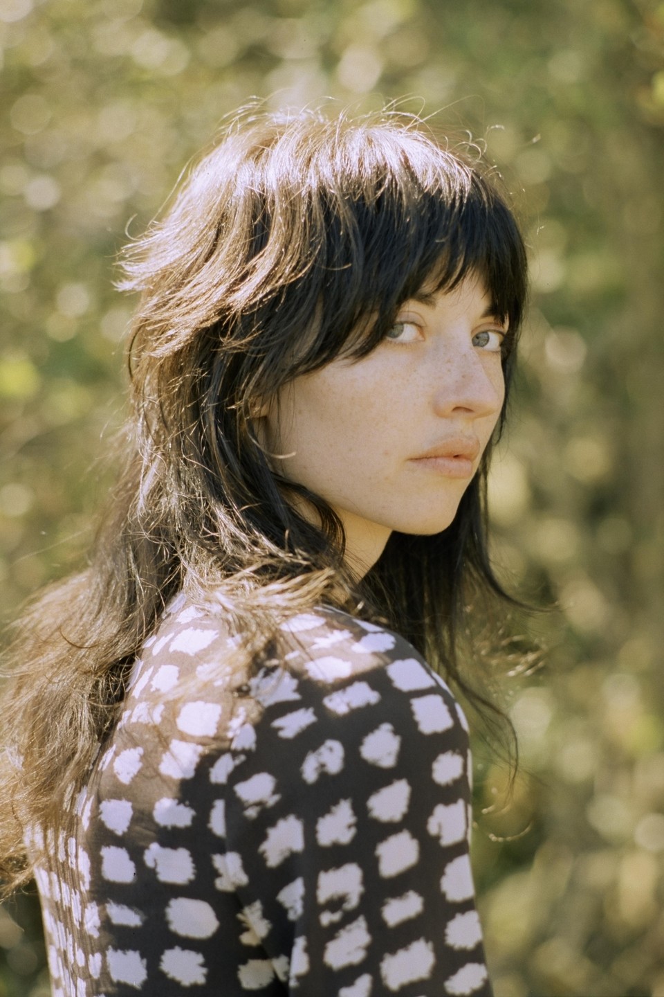 A woman with dark hair and bangs looks over her shoulder at the viewer.