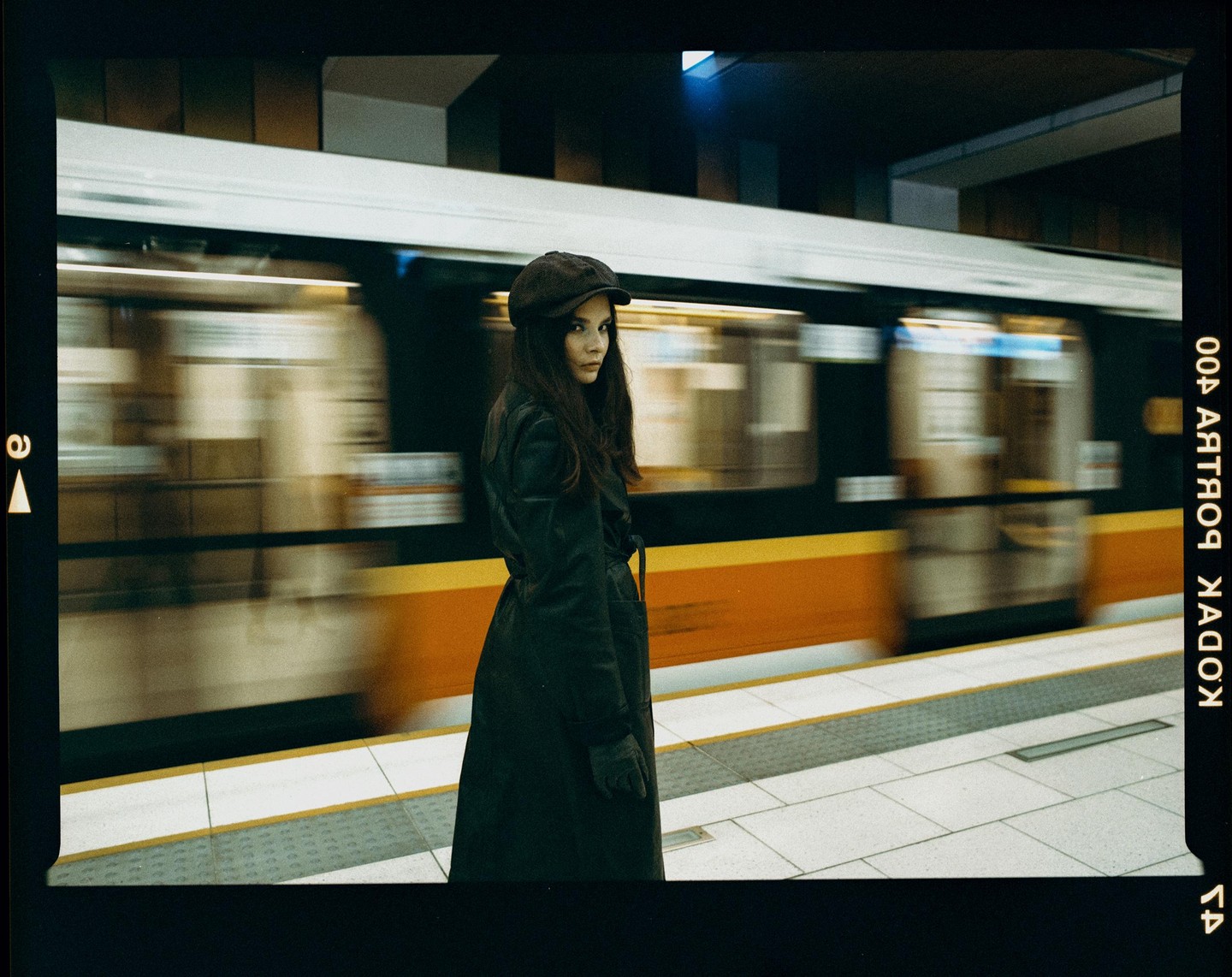 A woman in a coat and cap stands on a subway platform as a train passes by in a blur.