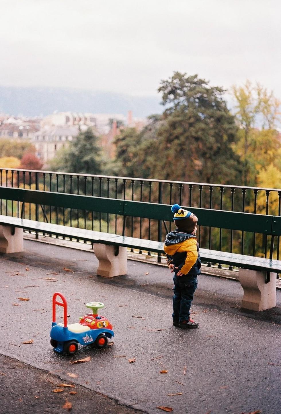 A child stands next to a toy car on a paved path overlooking a city.