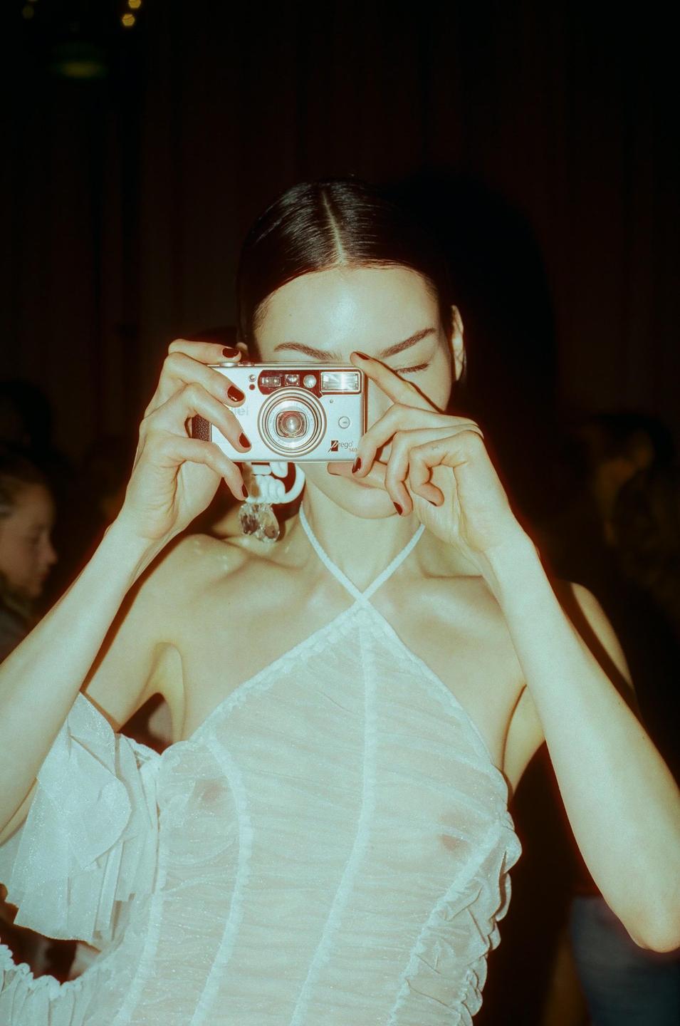 A woman in a white dress holds a silver point-and-shoot camera up to her face, obscuring her eyes.
