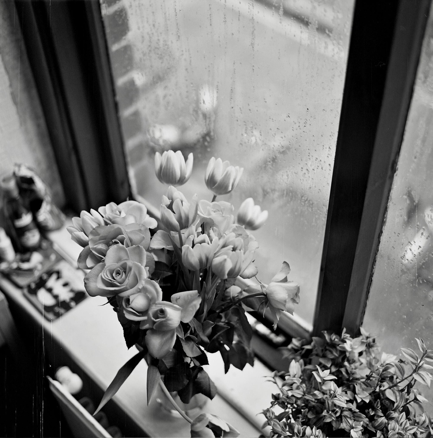 A bouquet of roses and tulips sits on a windowsill next to a rain-streaked window.