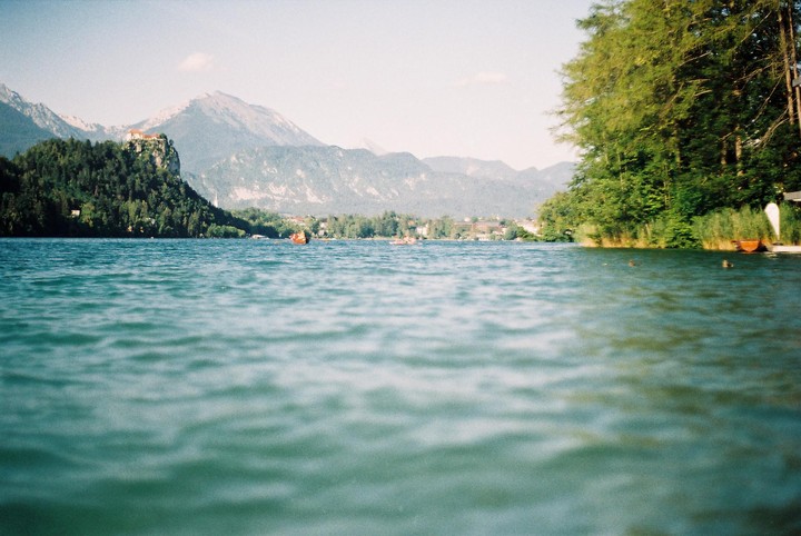 A lake with mountains and a castle on a hill in the background, and trees and boats on the shore.
