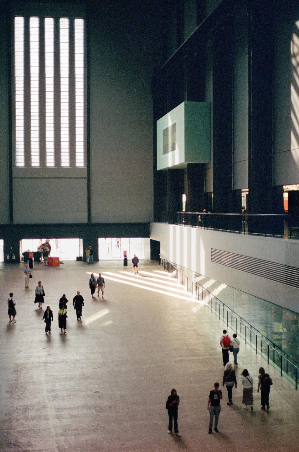 People walk across a large, open hall with tall windows and a balcony.