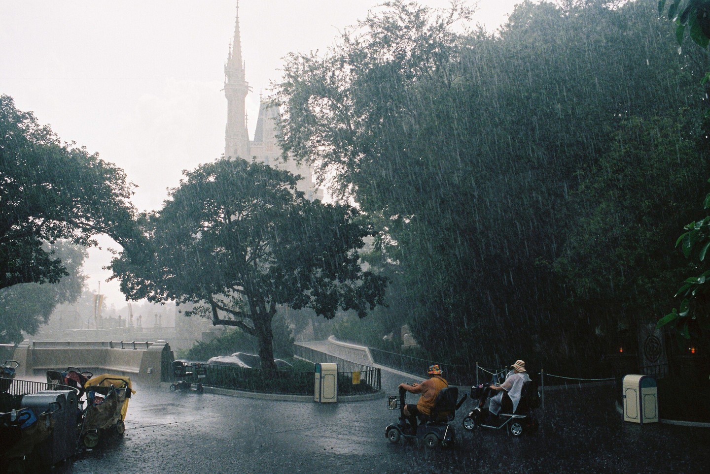 Two people on mobility scooters ride through a rainy street with a castle in the background.