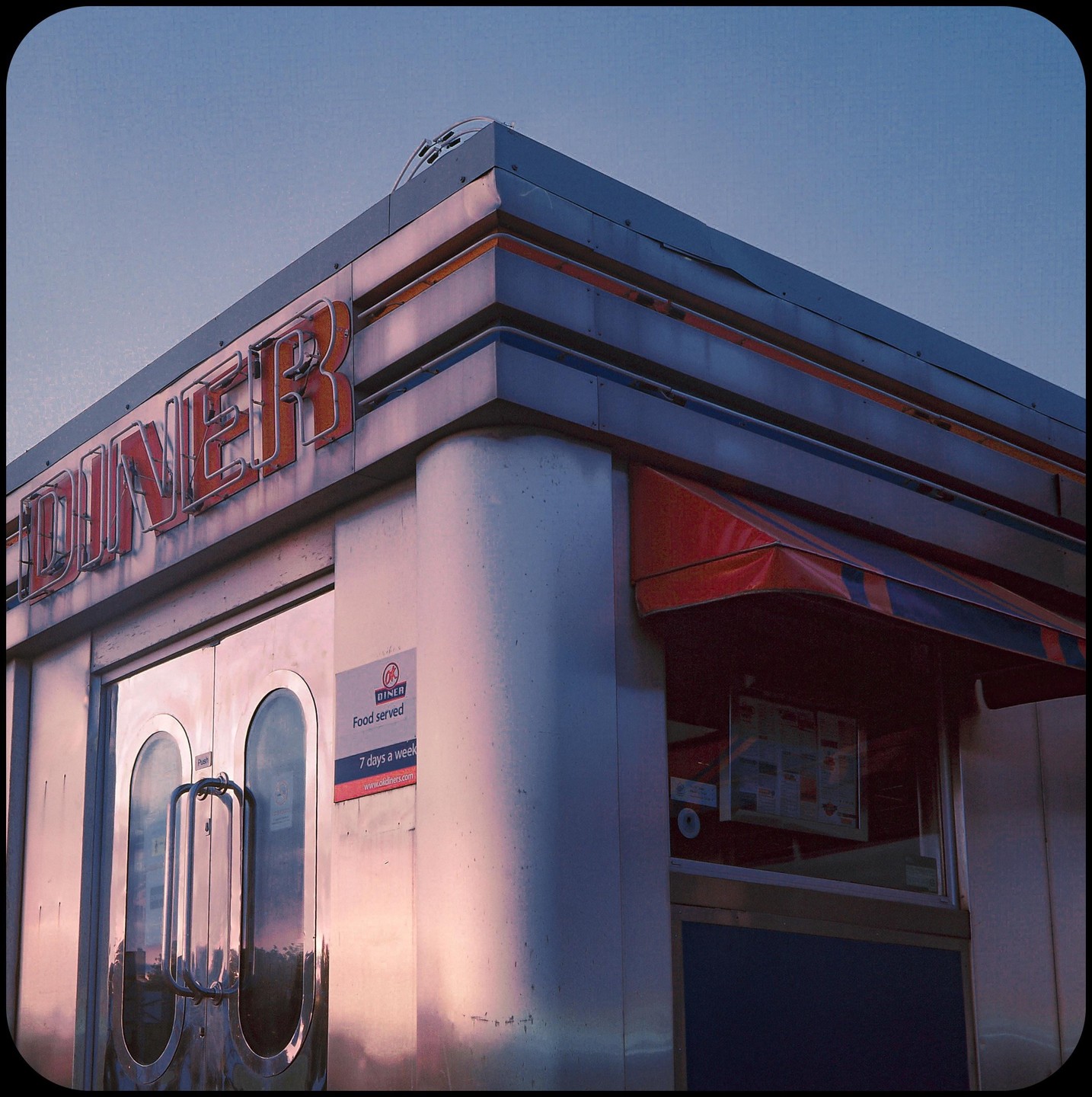 The exterior of a diner with a red awning is illuminated by the setting sun.