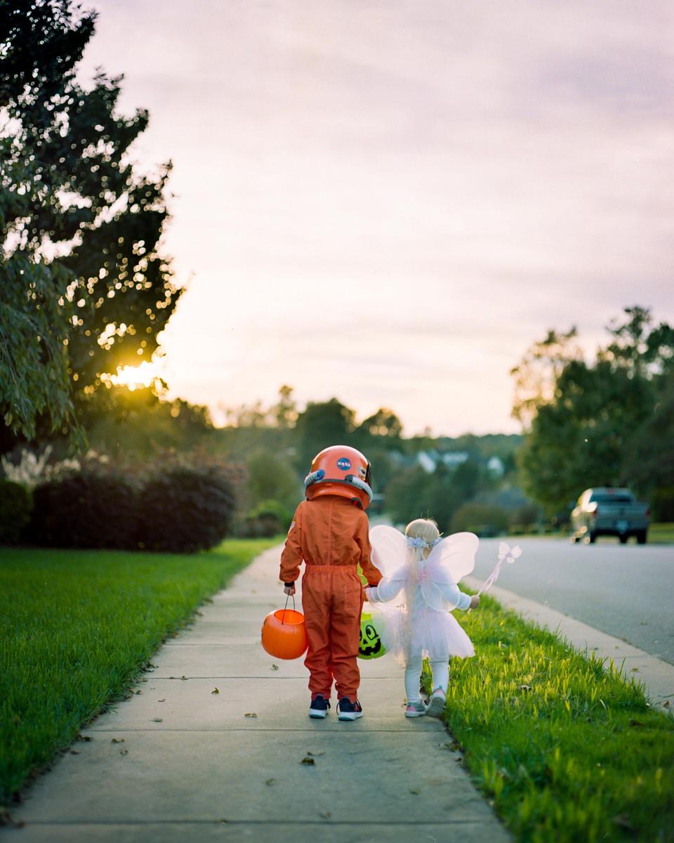 Two children in Halloween costumes walk along a sidewalk at sunset.