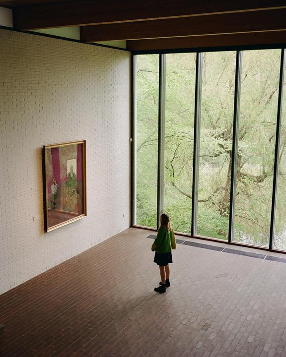 A woman stands in a large room with a painting on one wall and tall windows looking out onto trees.