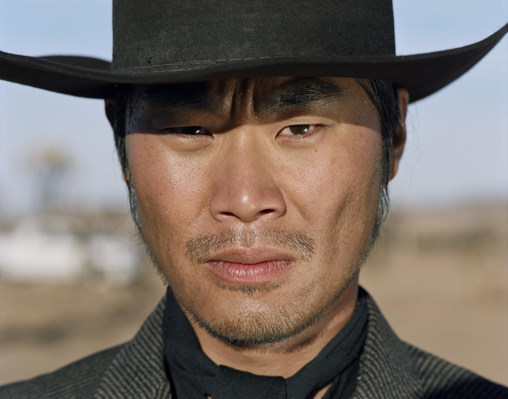 A man wearing a cowboy hat and a dark scarf looks directly at the viewer.