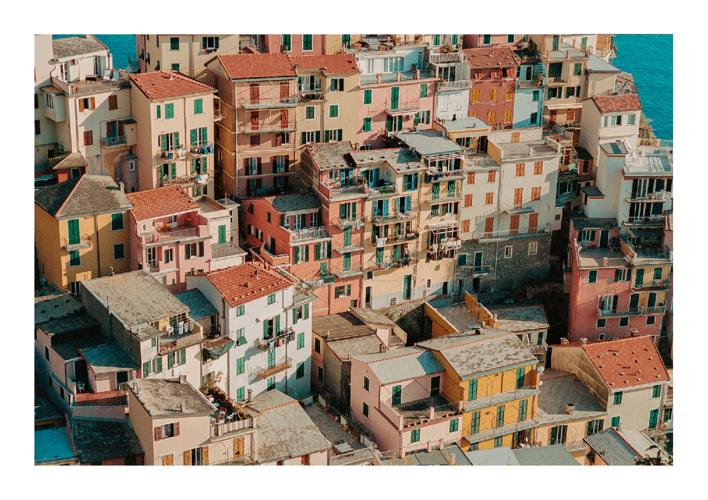 A high-angle view shows a cluster of colorful buildings with terracotta roofs nestled along a coastline.