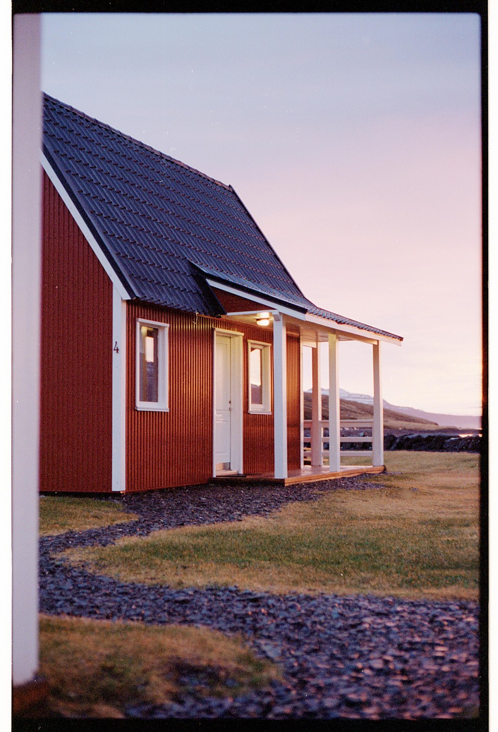 A red house with a porch and a dark roof stands on a grassy field with a gravel path leading to its entrance, under a pink and blue sky.