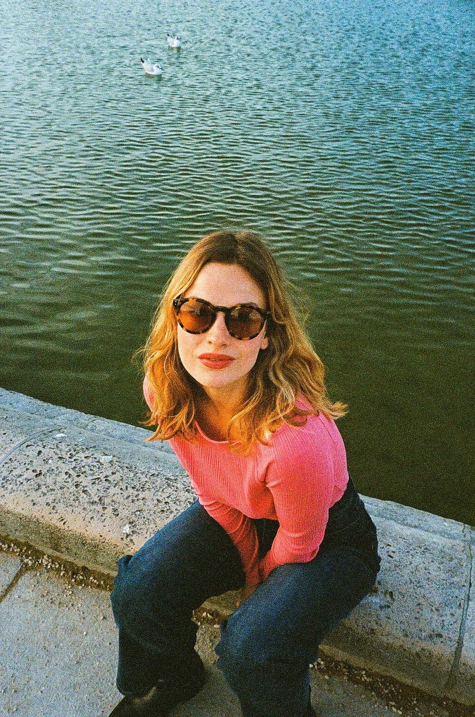 A woman wearing sunglasses and a pink shirt sits by the water with two birds in the background.