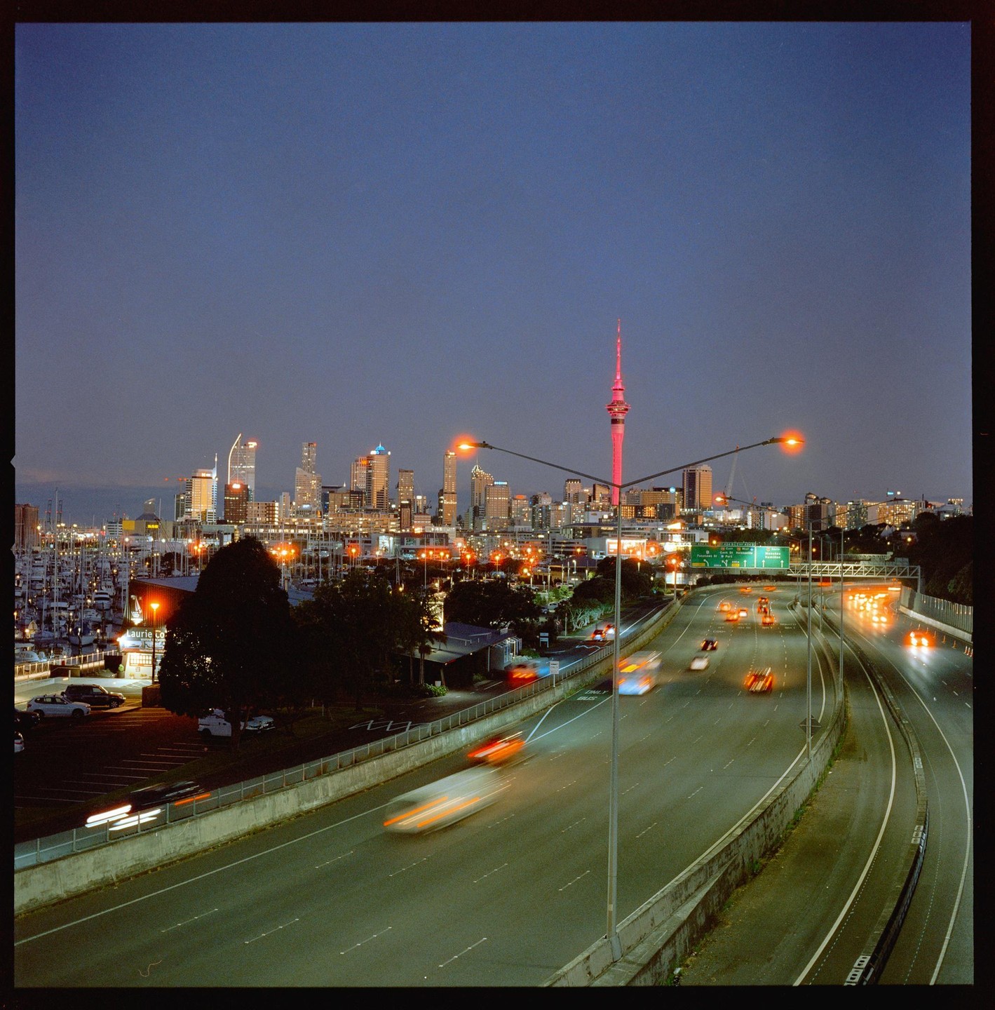 A highway with blurred car lights leads towards a city skyline with a prominent red tower and a marina at dusk.
