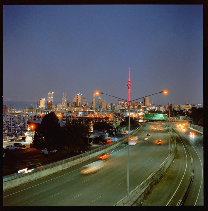A highway with blurred car lights leads towards a city skyline with a prominent red tower and a marina at dusk.