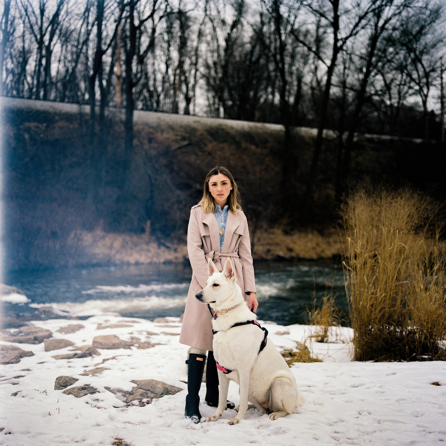 A woman and a white dog stand in the snow next to a river.