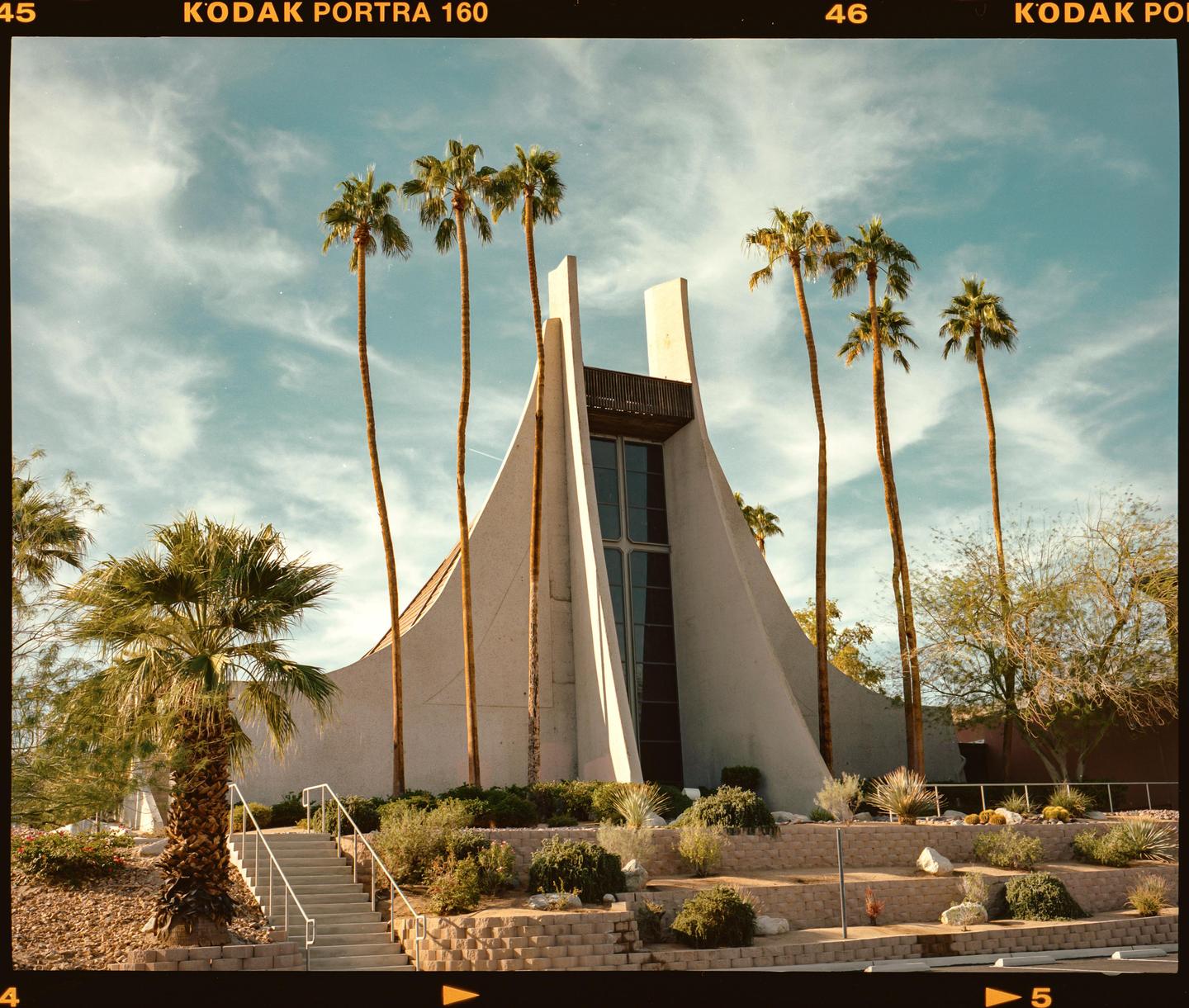 A modern building with a tall, pointed roof stands among palm trees and desert landscaping under a blue sky.