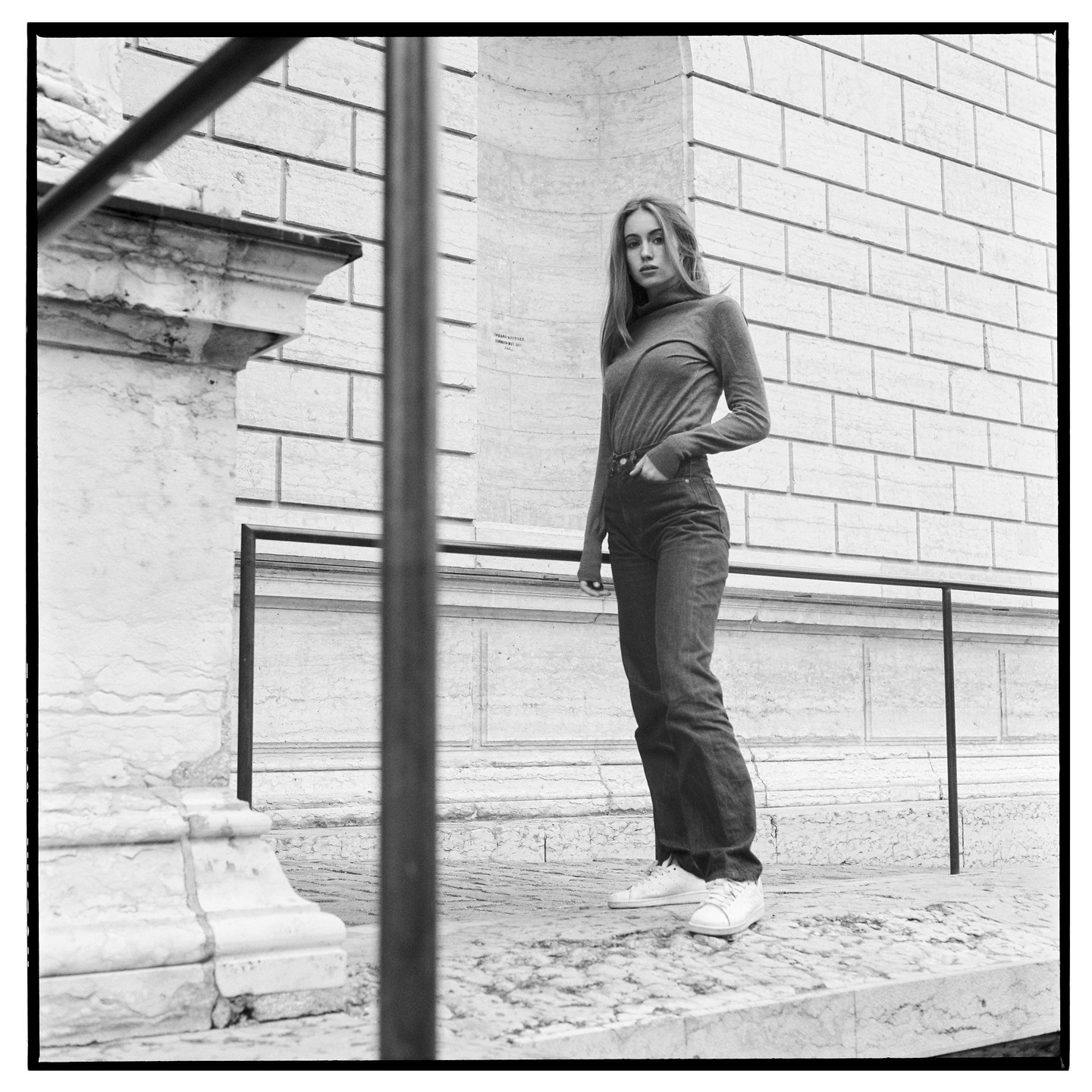 A woman stands on a stone ledge next to a building with a hand in her pocket.