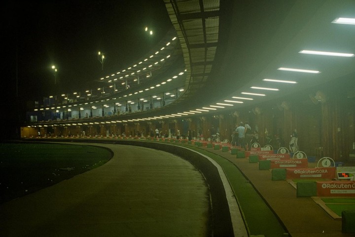 A multi-story golf driving range is illuminated at night, with several people visible on the lower levels.