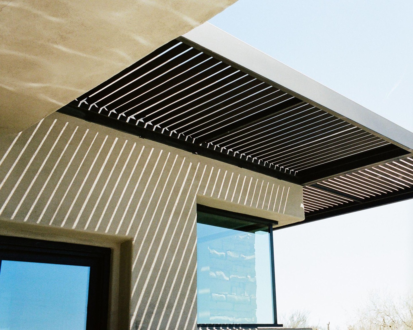 Sunlight casts diagonal shadows on the adobe wall of a building with a slatted overhang.