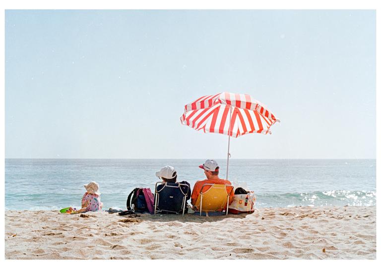 A family sits on a sandy beach under a red and white striped umbrella, looking out at the ocean.