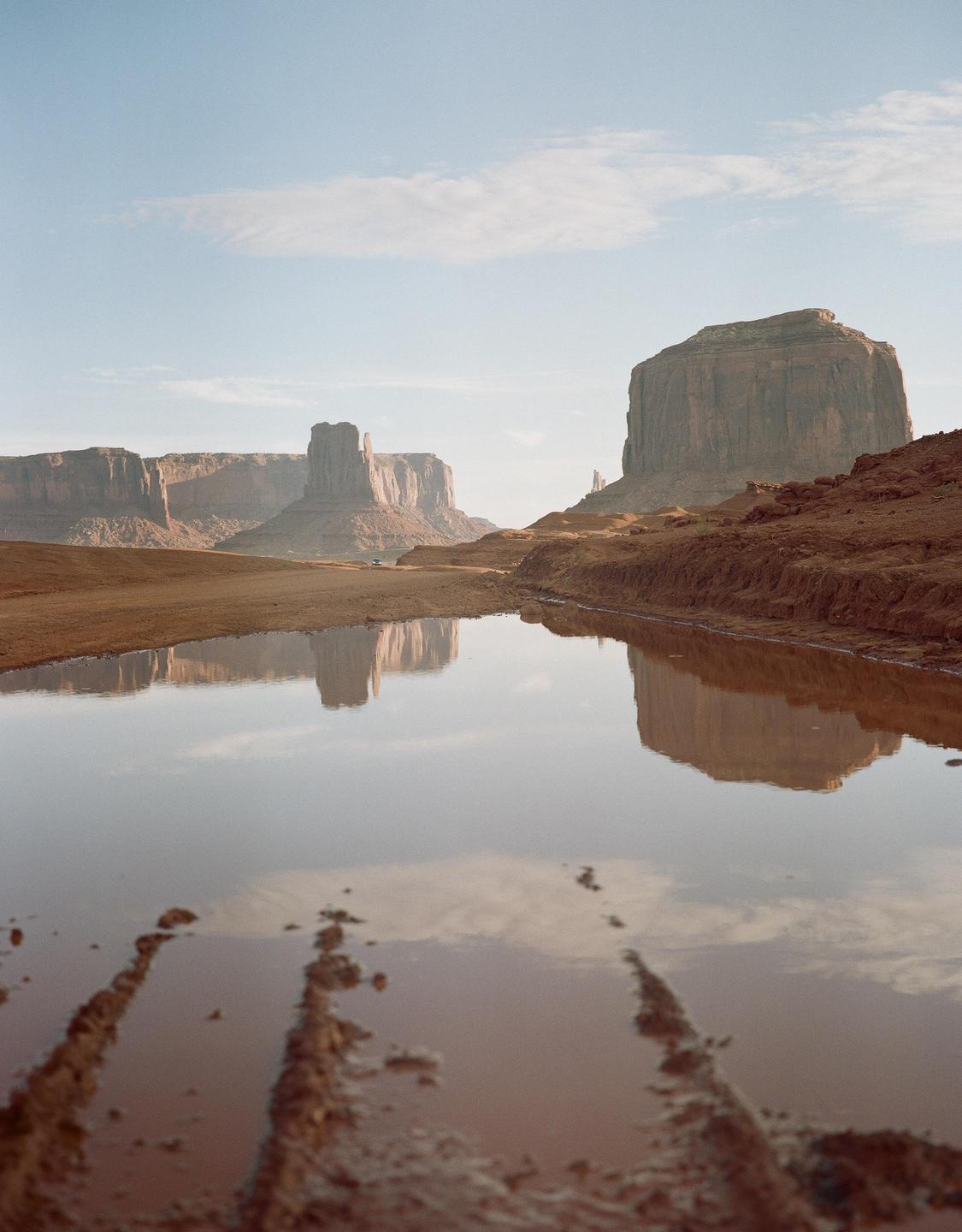A wide shot shows Monument Valley with a large puddle in the foreground reflecting the sky and the buttes.