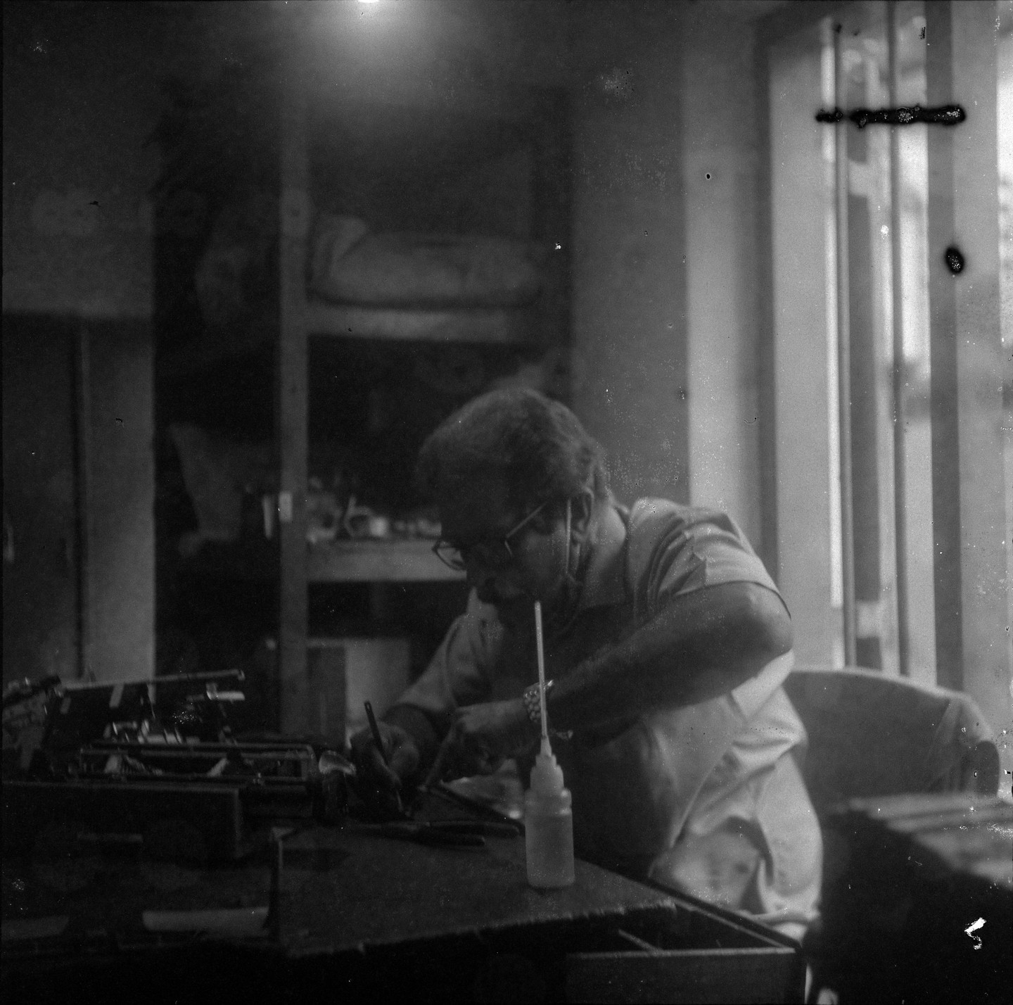 A man wearing glasses works on a typewriter at a desk next to a window.